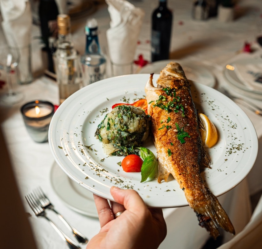 Prato com um peixe inteiro grelhado guarnecido com ervas, uma bola de purê de batatas com folhas verdes, um tomate cereja e uma rodela de limão, do restaurante Jadran em Budva, Montenegro.