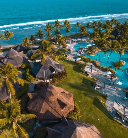 Vista aérea do Summerville Resort - All Inclusive à beira-mar com edifícios com telhados de palha, palmeiras, uma piscina e um litoral arenoso. Esse é um dos melhores resorts all inclusive no Brasil.
