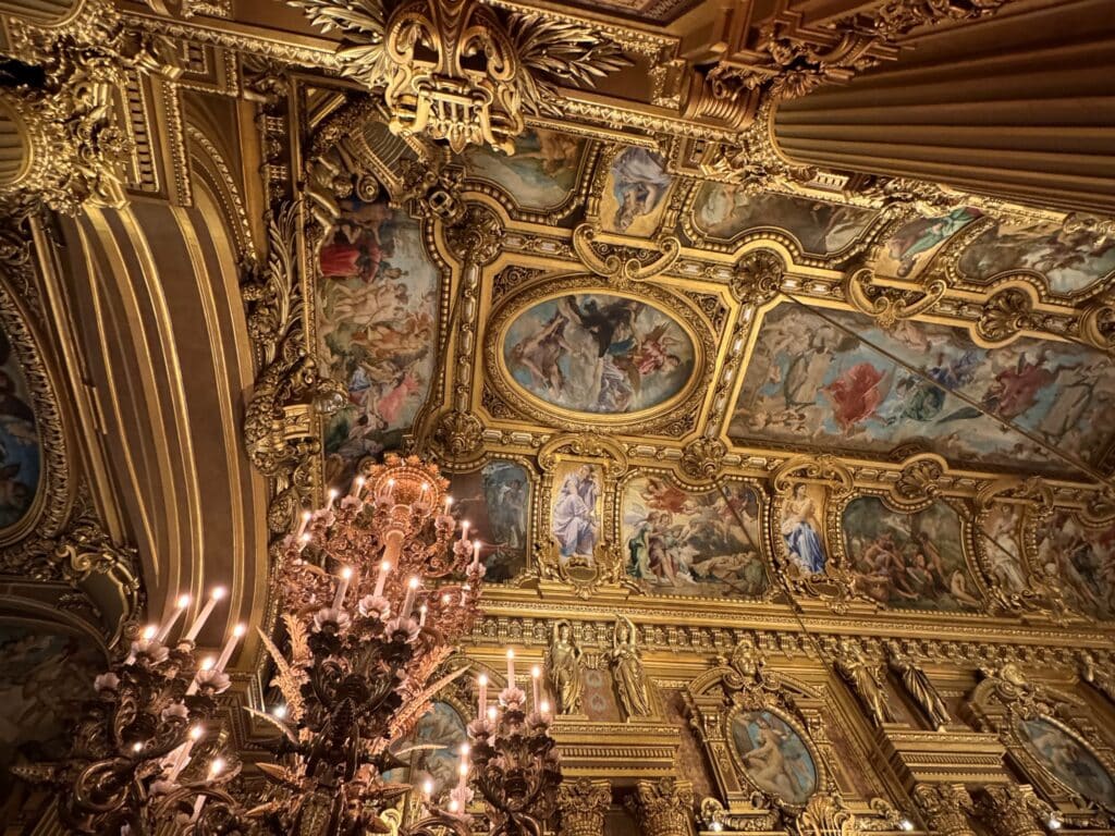 Teto dourado elaborado com afrescos detalhados e um lustre ornamentado em um espaço interior luxuoso. Esse é um dos pontos turísticos de Paris.