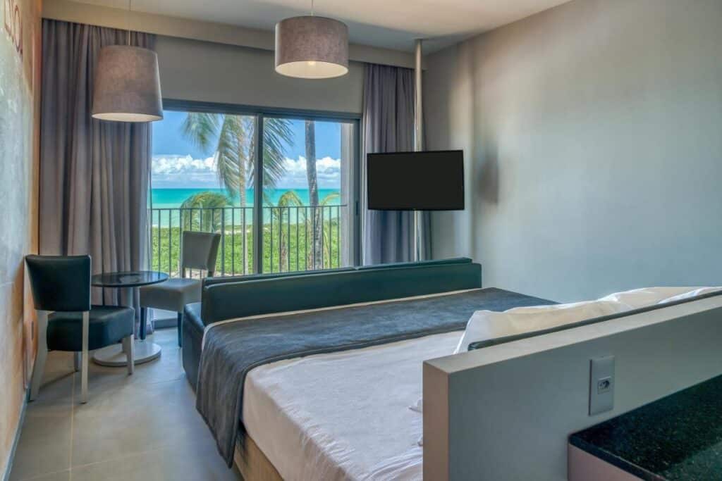 Um quarto da Vila Galé Resort Alagoas - All Inclusive com uma cama, duas cadeiras, uma mesa, uma TV e uma varanda com vista para as palmeiras e o oceano.