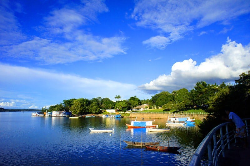 Uma vista tranquila do rio com pequenos barcos flutuando, cercada por vegetação exuberante e um céu azul parcialmente nublado.