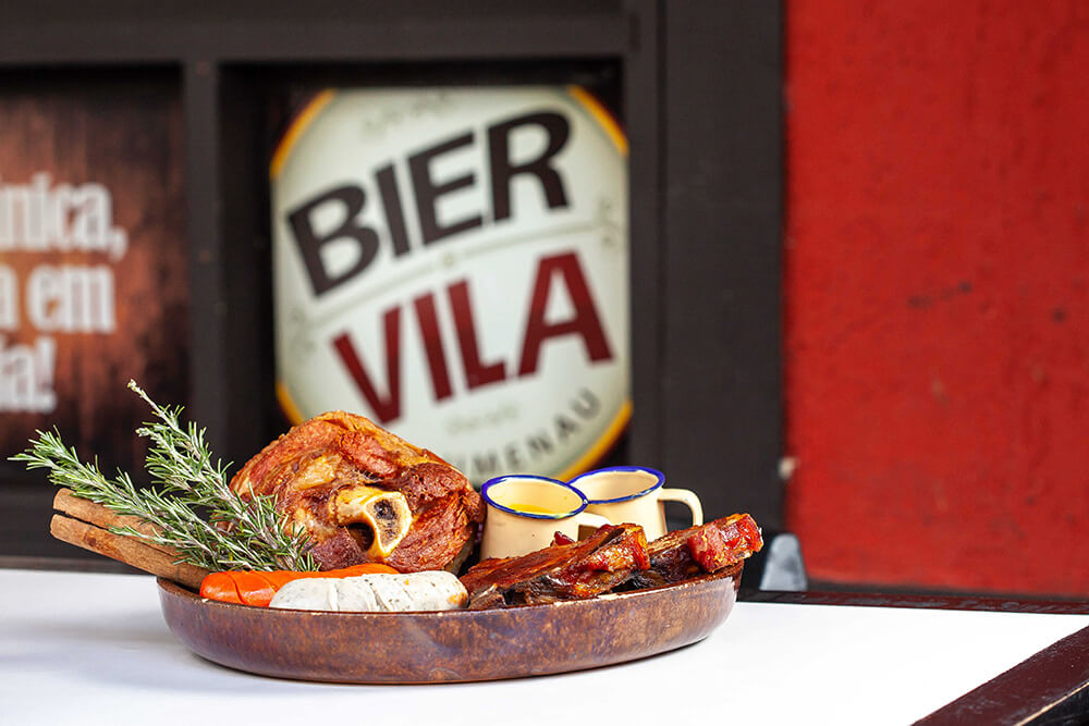 No Bier Vila, uma travessa de madeira com carne assada, salsichas e raminhos de alecrim. Duas canecas pequenas estão ao lado.