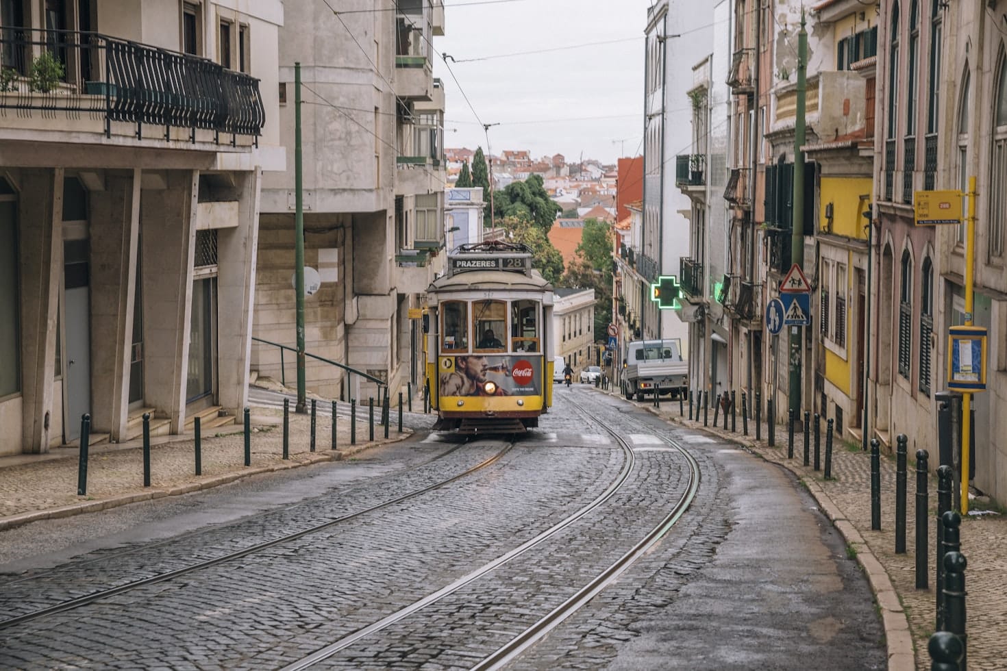 Um bonde amarelo na Calçada da Estrela, Lisboa viaja por uma rua de paralelepípedos em uma cidade, cercada por prédios. Há uma placa verde de farmácia à direita, sendo uma opção de roteiro para Portugal