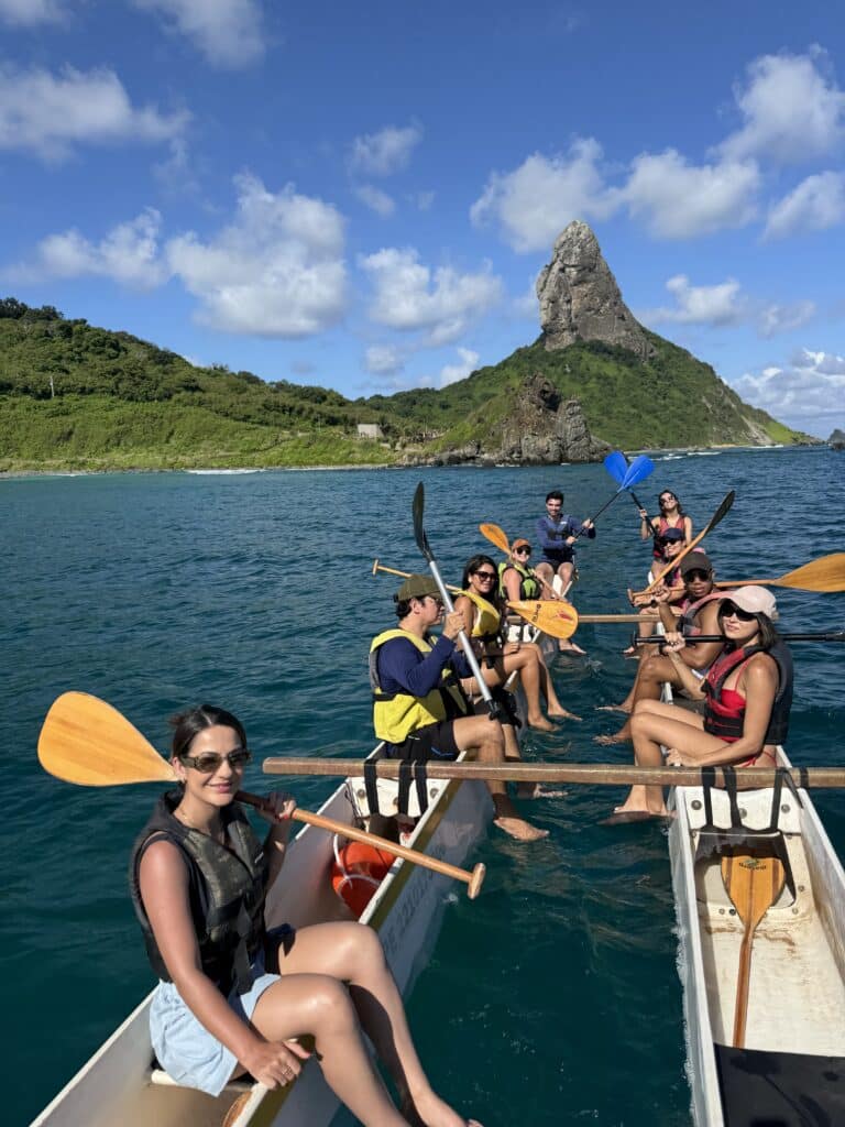 Dois grupos de pessoas em canoas remam em águas azuis perto de uma ilha verde com um alto pico rochoso ao fundo.