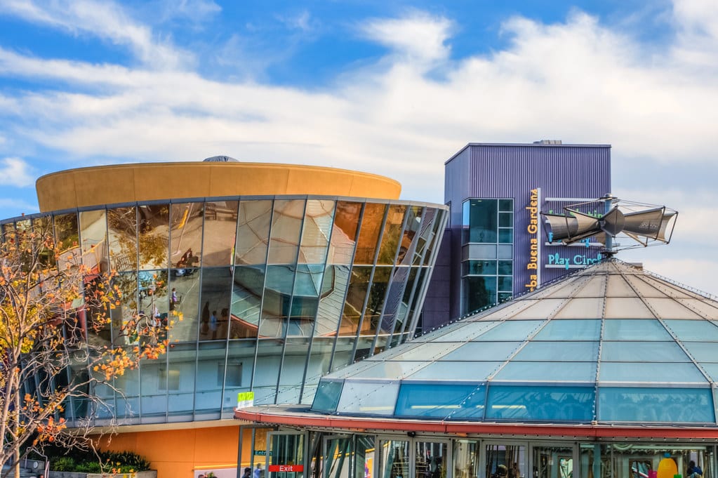 Edifício do Children’s Creativity Museum moderno com grandes janelas de vidro refletivo sob um céu azul com nuvens dispersas. Imagem para ilustrar post sobre museus em San Francisco.