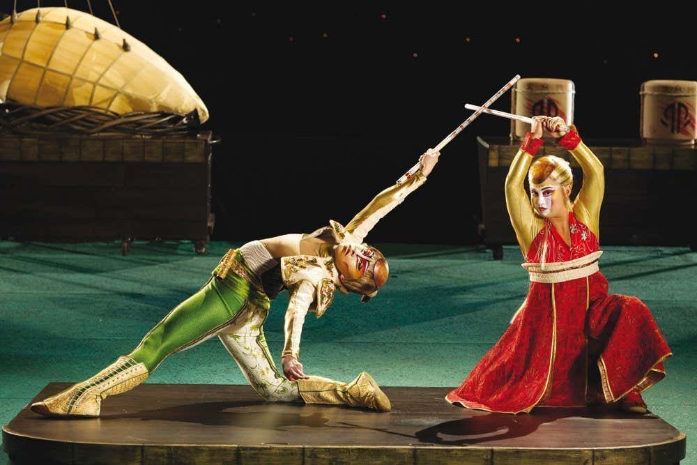 Dois artistas do do espetáculo KÁ", do Cirque du Soleil em Las Vegas. Eles estão em trajes elaborados e se envolvem em uma cena de luta coreografada com bastões no palco, cercados por adereços.