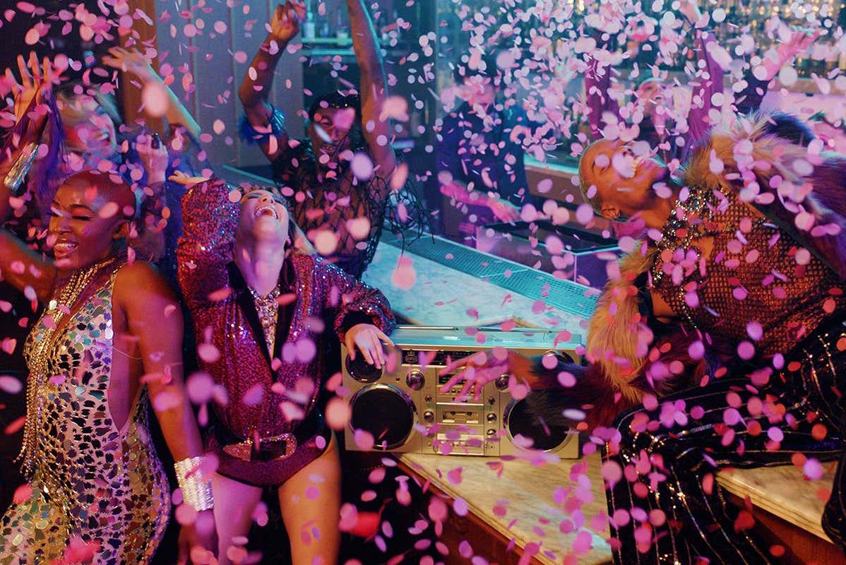 Grupo de pessoas do espetáculo Mad Apple, do Cirque du Soleil em Las Vegas. Eles estão em trajes coloridos dançando alegremente com confetes caindo, ao redor de um aparelho de som sobre uma mesa em um ambiente festivo.