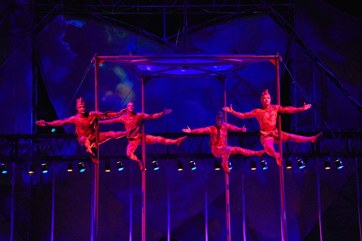 Quatro trapezistas do espetáculo Mystère, no Cirque du Soleil em Las Vegas. Eles estão em trajes vermelhos e realizam movimentos sincronizados em postes verticais sob iluminação colorida do palco.