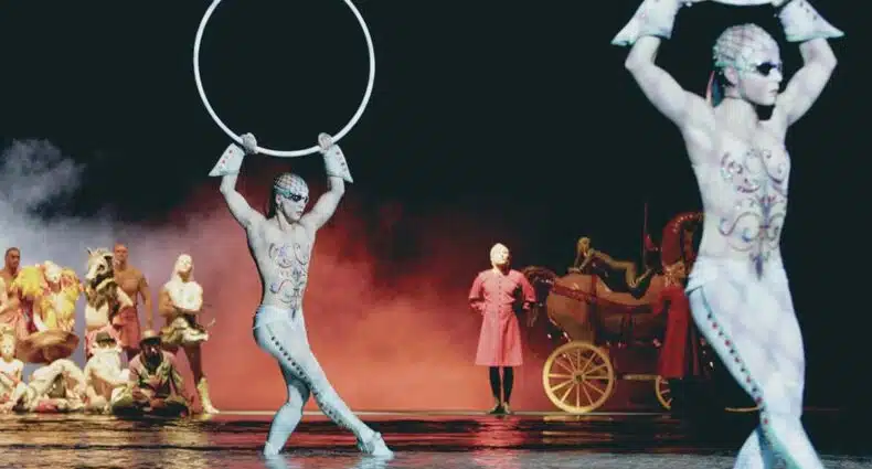 Artistas do espetáculo "O", do Cirque du Soleil em Las Vegas, em trajes estampados com anéis no palco, outros no fundo em trajes coloridos e uma carruagem vermelha.