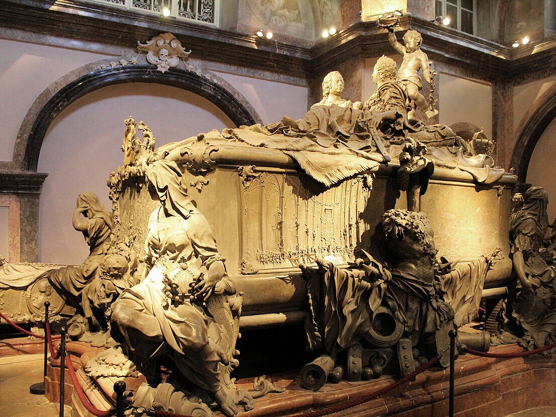 Sarcófago de pedra na Cripta Imperial. Ele é ornamentado com figuras esculpidas e cortinas em uma sala mal iluminada, cercada por barreiras de corda.