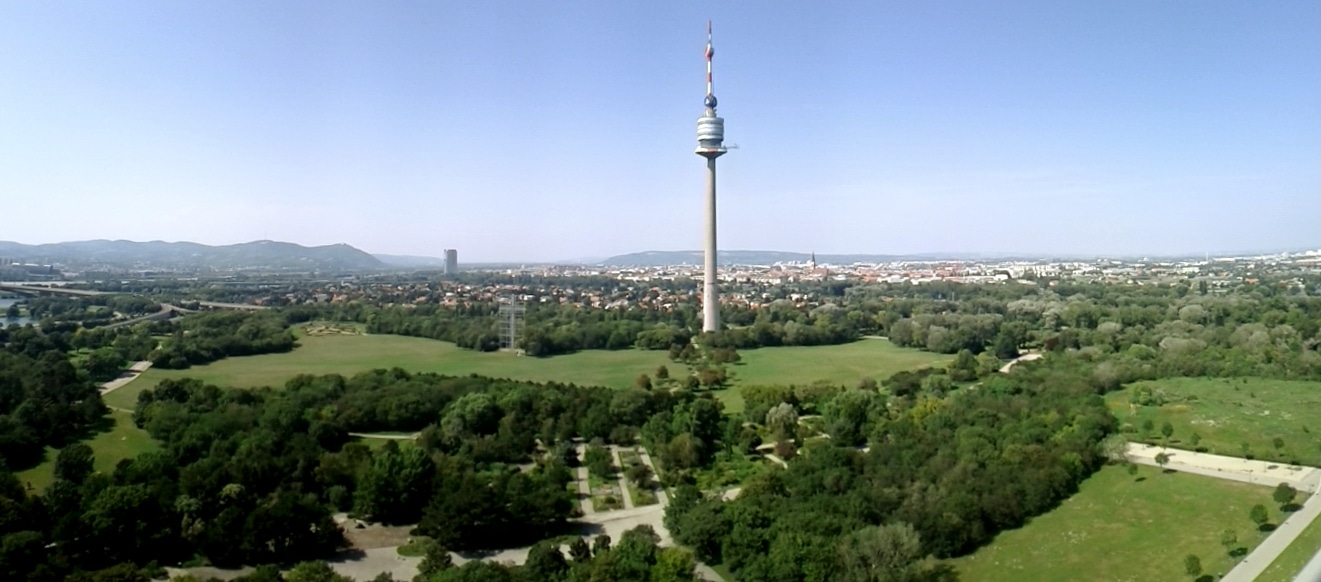 Vista panorâmica do Donaupark. A torre do Danúbio é alta fica em meio a um vasto parque verde com caminhos, árvores e uma paisagem urbana distante sob um céu azul claro.