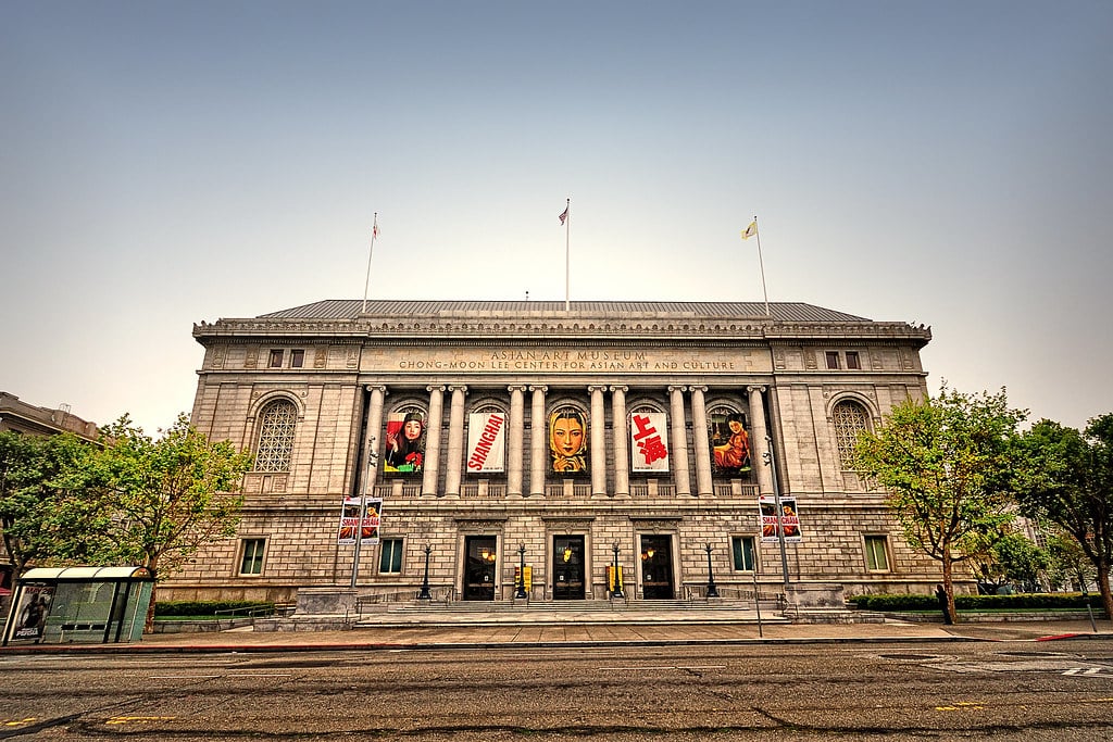 Asian Art Museum com faixas e cartazes em colunas, cercado por árvores e um ponto de ônibus em frente. Imagem para ilustrar post sobre museus em San Francisco.