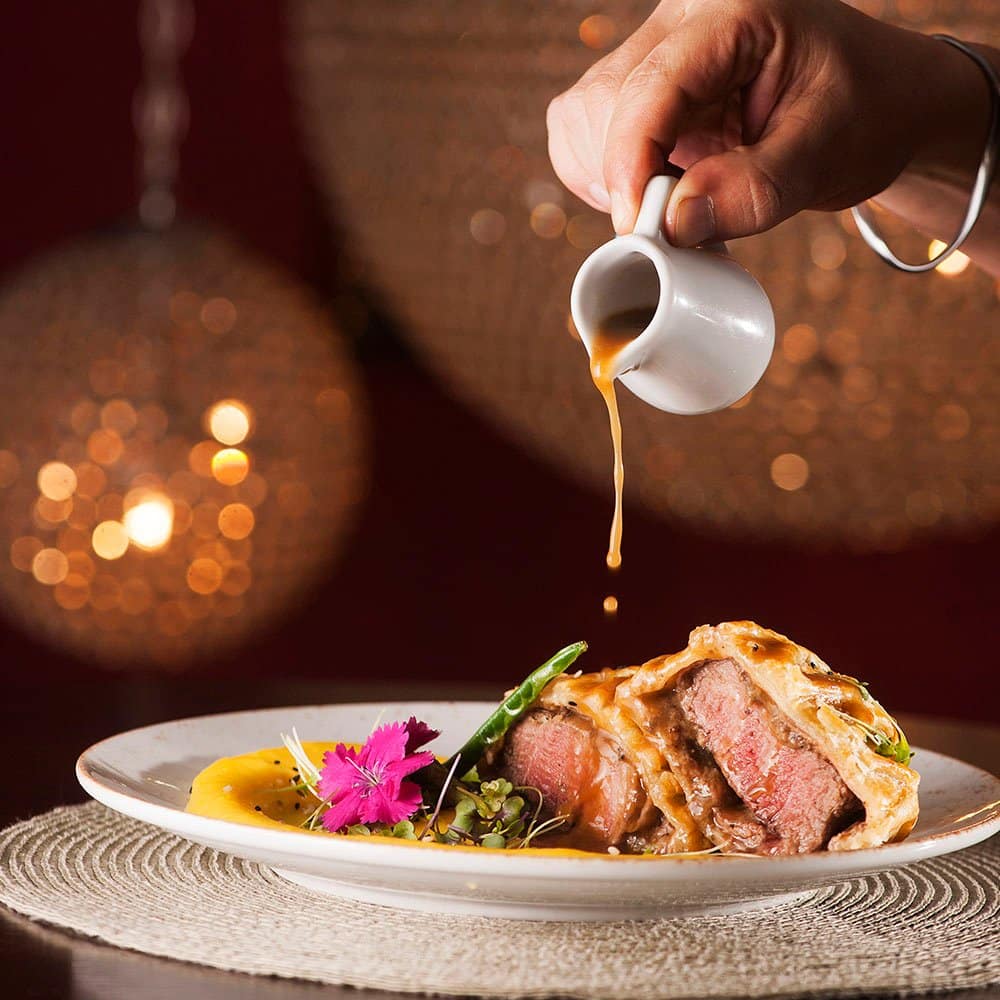 Mão despejando molho sobre um prato de carne com legumes e guarnição, contra um fundo de luz decorativa desfocado.