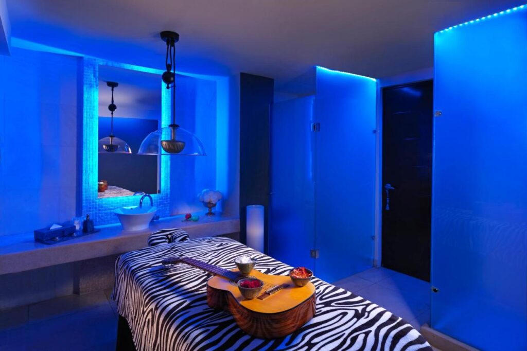 Uma sala do Hard Rock Hotel Cancun com iluminação azul, com uma mesa de massagem com capa estampada de zebra, um violão e uma pia com espelho.