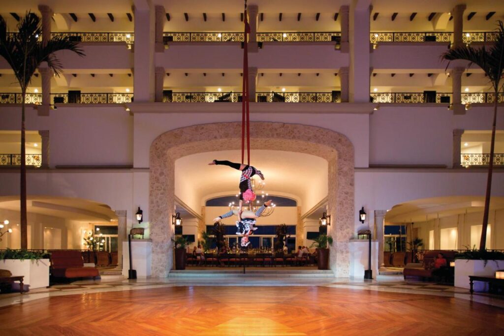 Dois acrobatas realizam uma rotina aérea em sedas vermelhas em um grande e elegante saguão do Hyatt Zilara Cancun com varandas e iluminação decorativa.