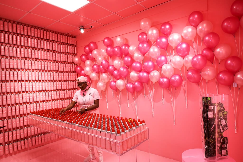 Uma pessoa está em uma sala cheia de paredes rosas, balões rosas e brancos e uma mesa coberta com latas de tinta spray vermelha do Ice Cream Museum.