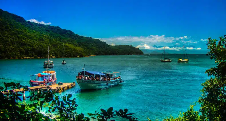 Um cenário costeiro vibrante com barcos no cais e vários veleiros nas águas azul-turquesa, cercados por colinas verdejantes.