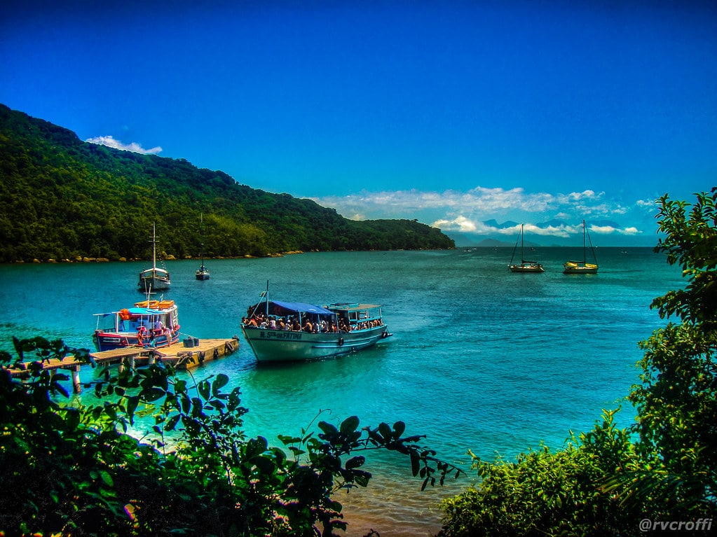 Um cenário costeiro vibrante com barcos no cais e vários veleiros nas águas azul-turquesa, cercados por colinas verdejantes.