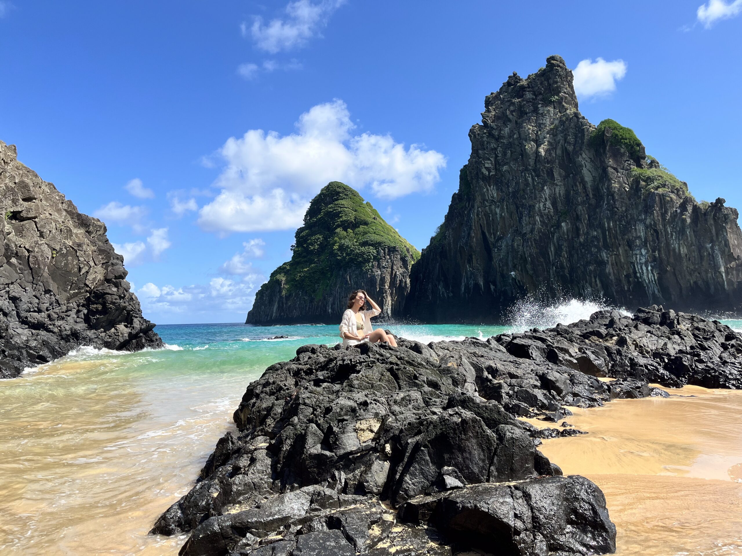 Uma pessoa sentada em rochas negras perto do oceano, com grandes ilhas rochosas e céu azul ao fundo.
