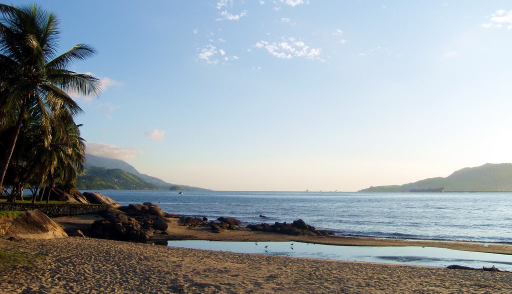 Uma cena de praia serena com águas calmas, montanhas distantes, palmeiras e uma pequena área de areia sob um céu azul claro. Essa é a praia do do Itaguaçu em Ilhabela, um dos melhores lugares para curtir o Réveillon no Brasil.
