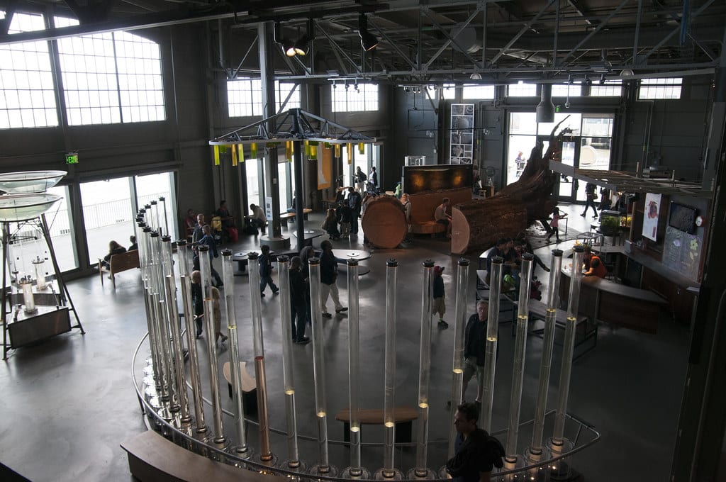 Interior do Exploratorium com grandes janelas, vigas de metal, exposições interativas e pessoas caminhando e interagindo com as exibições.