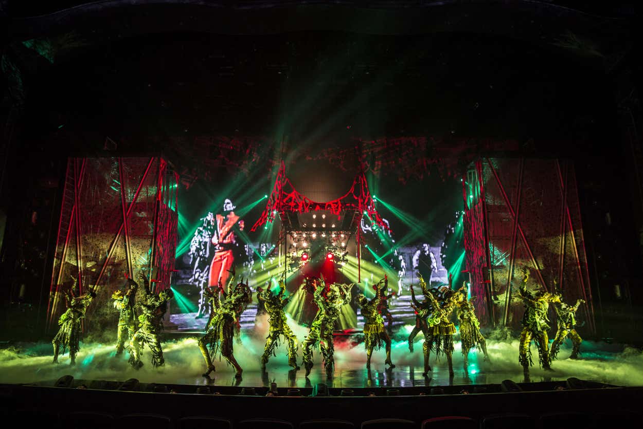 Dançarinos do espetáculo Michael Jackson One fantasiados se apresentam energicamente em um palco cheio de neblina, com luzes vermelhas e verdes vibrantes e grandes imagens projetadas.
