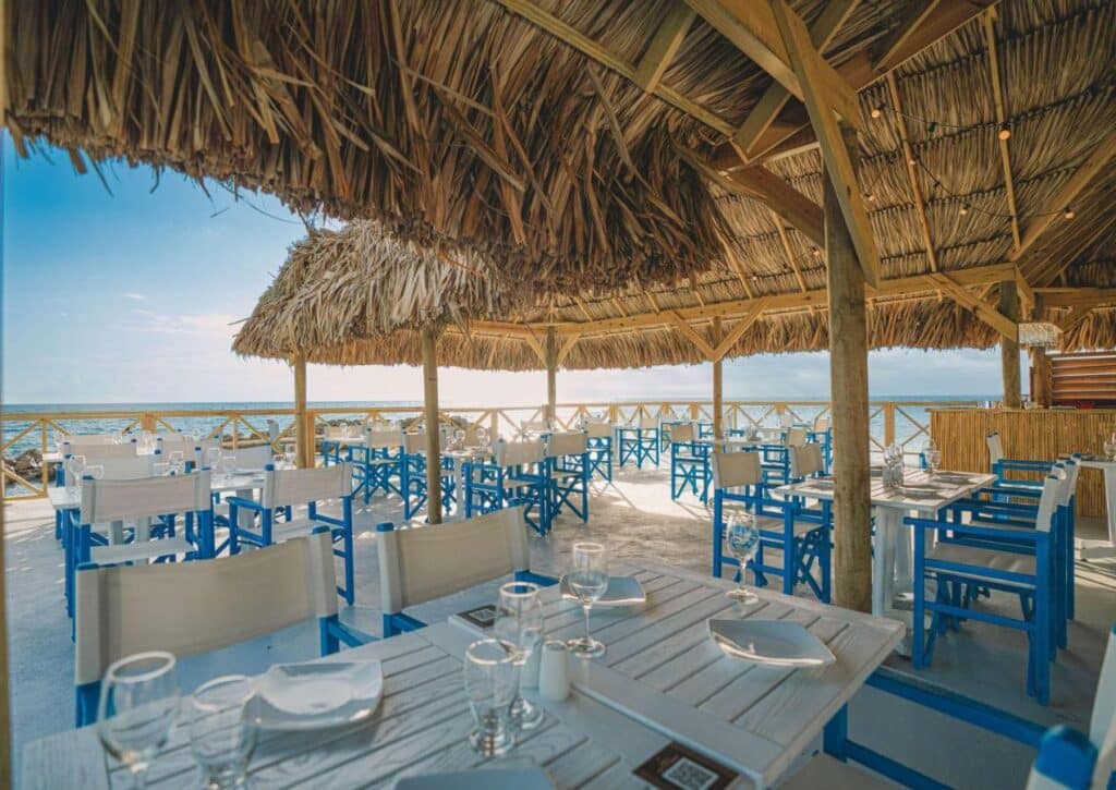 Restaurante ao ar livre à beira-mar do Mangrove Beach Corendon com mesas de madeira, cadeiras brancas e telhado de palha, com vista para o oceano em um dia ensolarado. Esse é um dos melhores resorts all inclusive no Caribe.