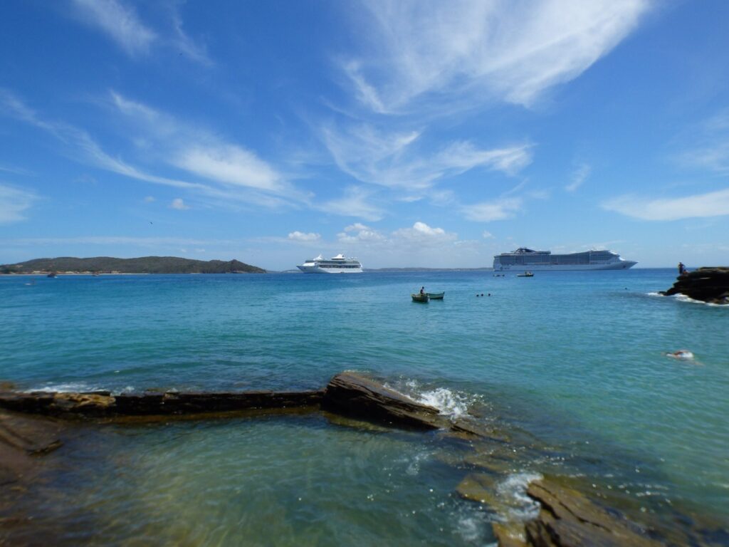 Vista do oceano com dois navios de cruzeiro ao longe, céu azul claro e uma costa rochosa em primeiro plano. Essa praia fica em Búzios, um dos melhores lugares para curtir o Réveillon no Brasil.