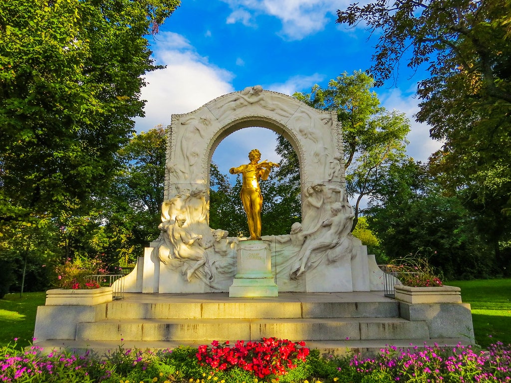 Monumento de Johann Strauss no Stadtpark. Se trata de uma estátua dourada de um violinista fica sob um arco decorativo com figuras esculpidas em um parque, cercado por árvores e canteiros de flores.