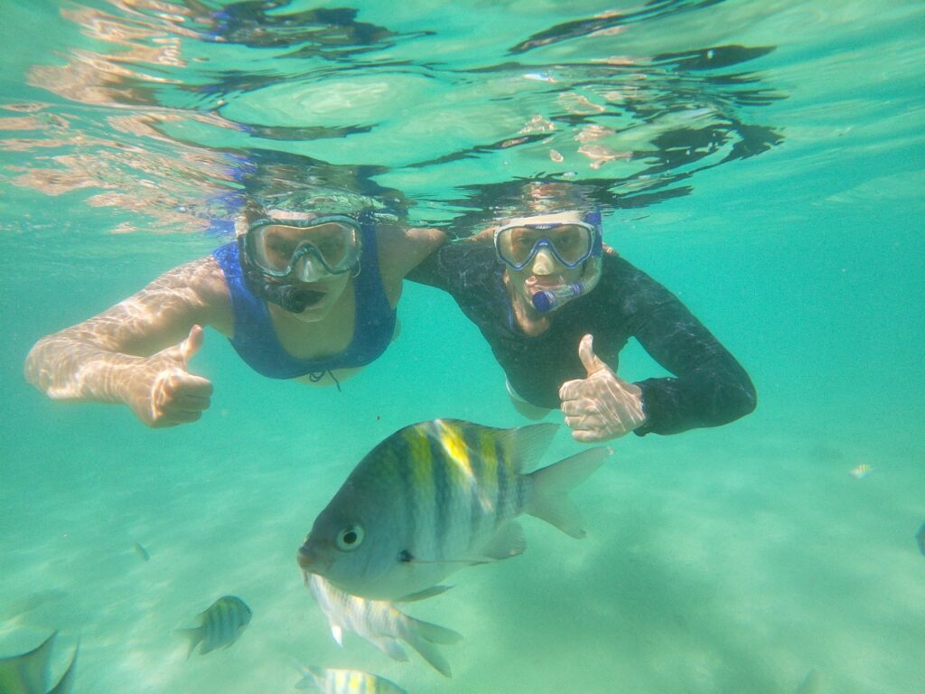 Dois mergulhadores debaixo d'água fazem sinal de positivo perto de um peixe tropical com listras amarelas e pretas em águas azul-turquesa cristalinas.