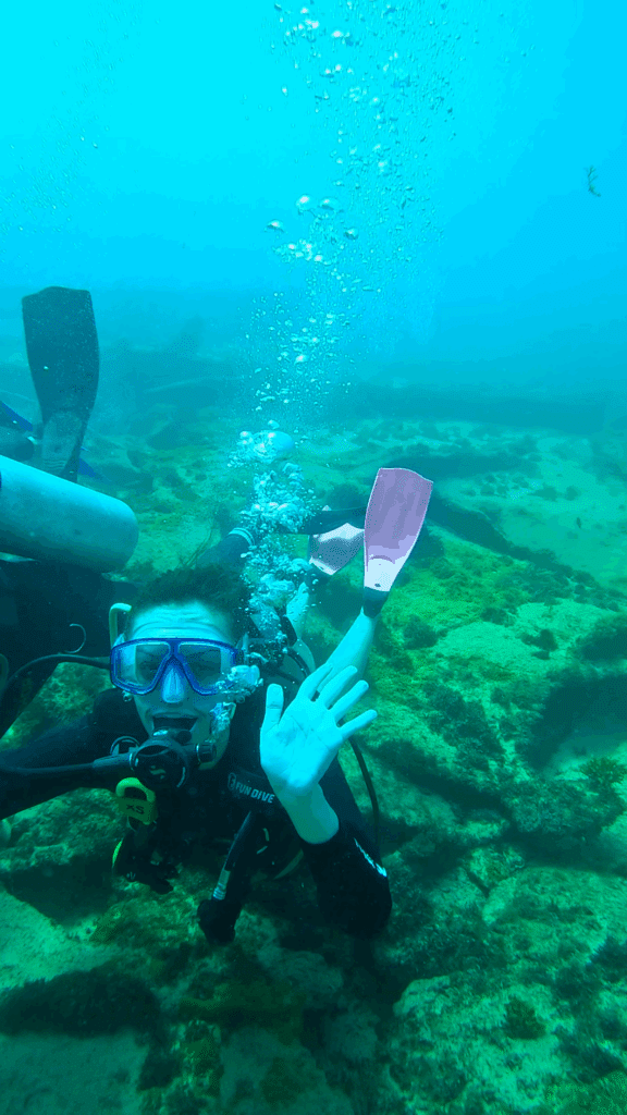 Um mergulhador usando máscara e equipamento acena para a câmera em um ambiente rochoso e esverdeado.
