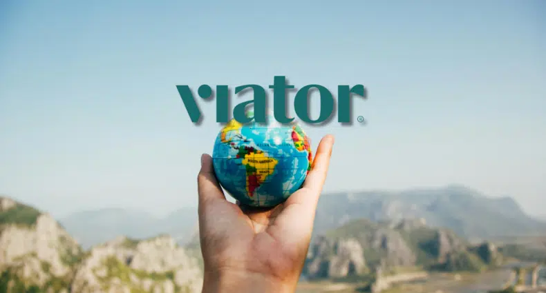 Uma pessoa segura um pequeno globo na mão com uma paisagem montanhosa ao fundo. A palavra "Viator" está sobreposta, para ilustrar o post de Viator é confiável