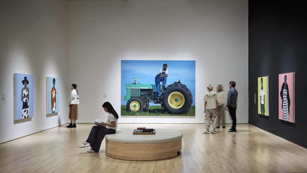 Museu de Arte Moderna de San Francisco apresenta uma grande foto de trator e pinturas de pessoas. Uma mulher está sentada em um banco, lendo. Outros observam a arte.