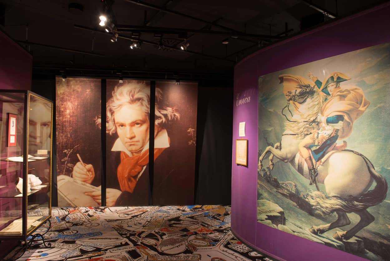 Exposição Museum Haus der Musik (Casa da Música) com grandes retratos de Beethoven e Napoleão, cercada por paredes roxas e um piso estampado. A foto ilustra o post de o que fazer em Viena.