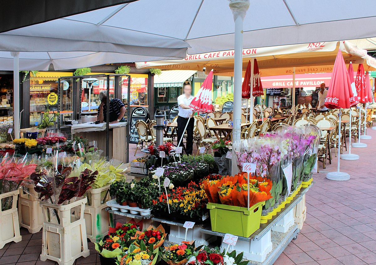 Foto do Naschmarkt. O mercado é ao ar livre com barracas de flores e um café ao fundo. Flores são exibidas em baldes sob grandes guarda-sóis brancos. A foto ilustra o post de o que fazer em Viena.
