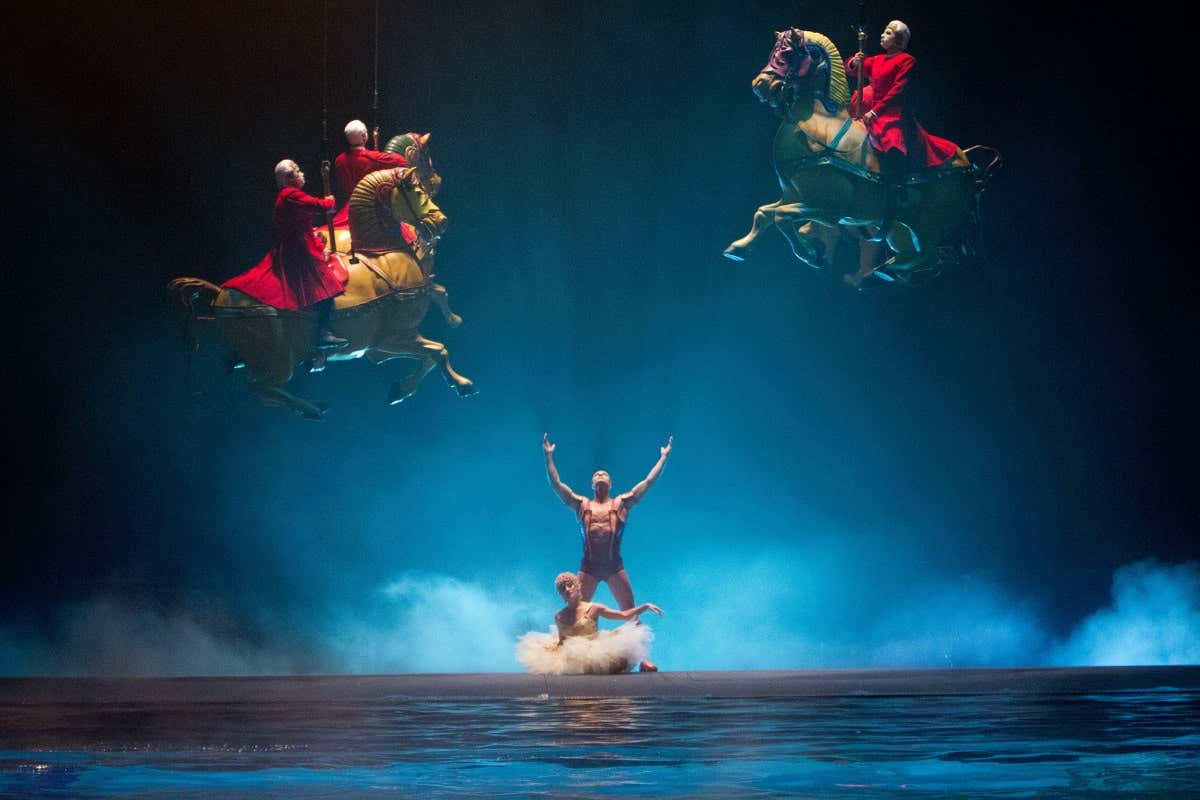 Artistas no palco; dois usam capas vermelhas e montam cavalos suspensos, enquanto um dançarino posa com os braços levantados sob um holofote azul.