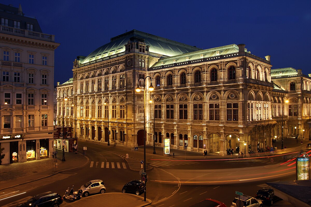 Ópera de Viena durante à noite. O edifício é histórico e ornamentado, com janelas em arco e telhado verde iluminado por postes de luz, em uma esquina movimentada.