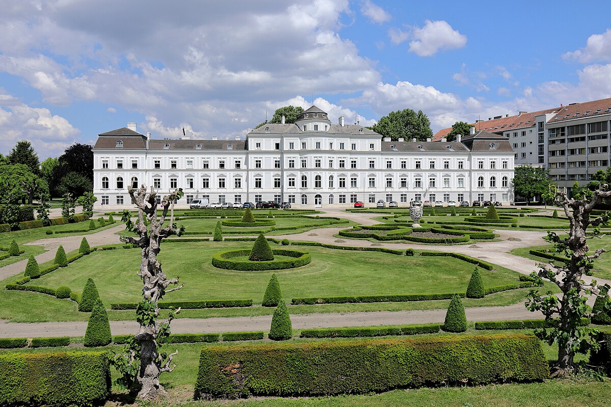 Vista do Palácio Augarten. Ele é todo branco com várias janelas, visto atrás de jardins bem cuidados com sebes e padrões geométricos sob um céu parcialmente nublado. A foto ilustra o post de o que fazer em Viena.