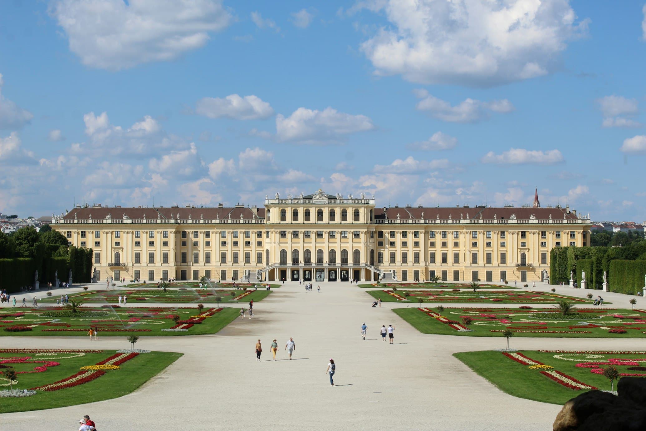 A imagem mostra o amplo Palácio de Schönbrunn com seus jardins e caminhos simétricos, sob um céu azul parcialmente nublado. É uma opçõa de o que fazer em Viena.