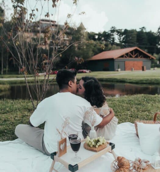 Um casal sentado sobre um cobertor branco, dando um beijo durante um piquenique perto de um lago, com uma bandeja de madeira com vinho e salgadinhos por perto.