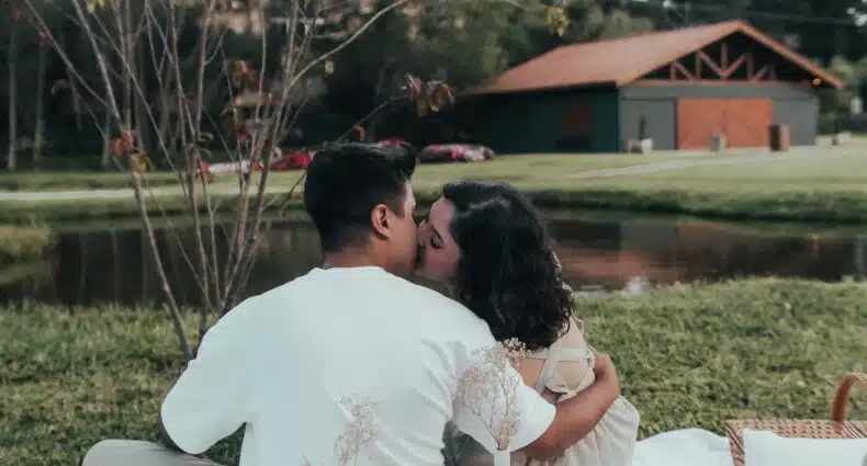 Um casal sentado sobre um cobertor branco, dando um beijo durante um piquenique perto de um lago, com uma bandeja de madeira com vinho e salgadinhos por perto.