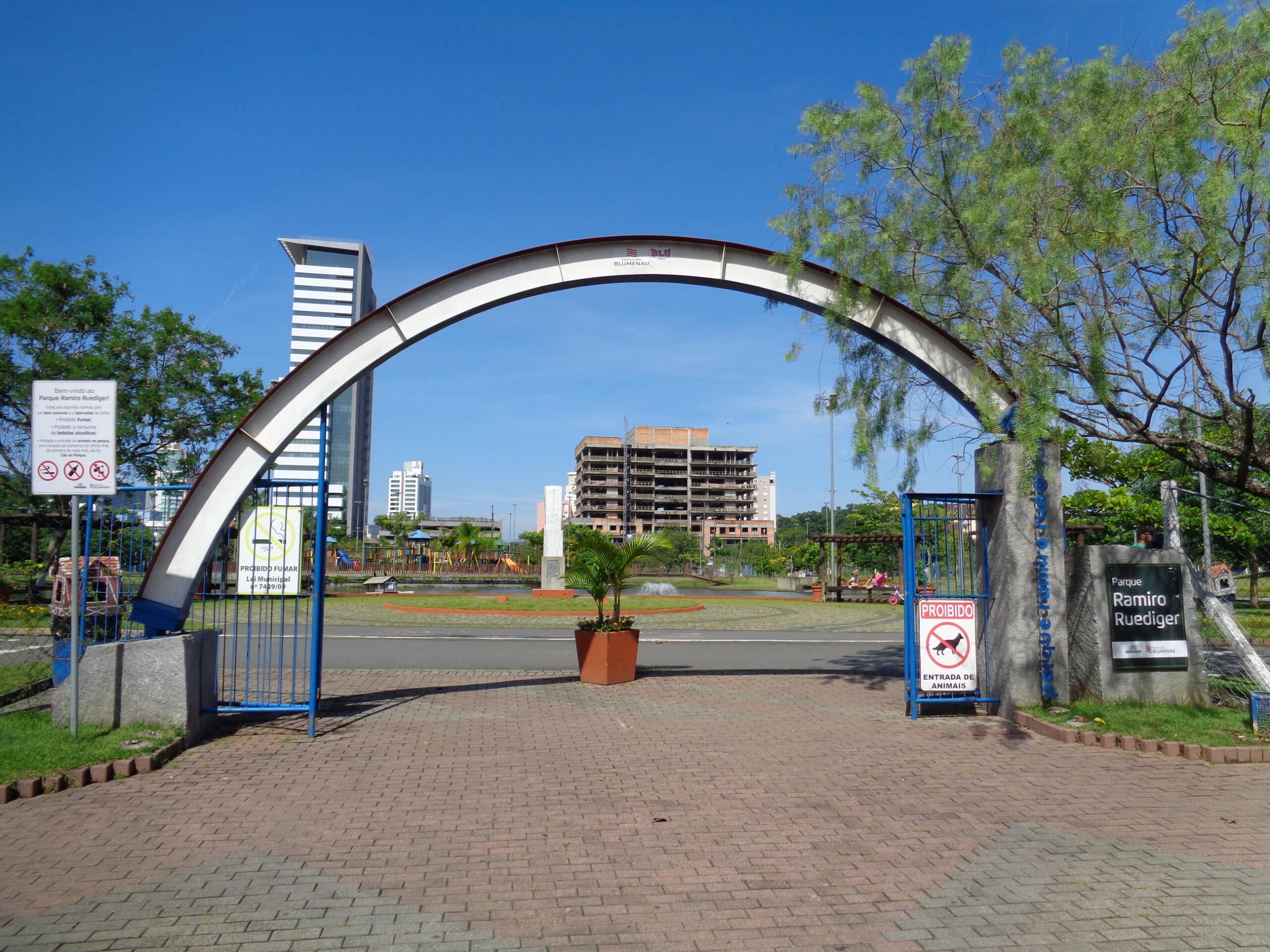 Entrada em arco para o Parque Ramiro Ruediger com caminho de tijolos, placas, árvores e uma paisagem urbana ao fundo sob um céu azul claro. Imagem para ilustrar post sobre o que fazer em Blumenau.