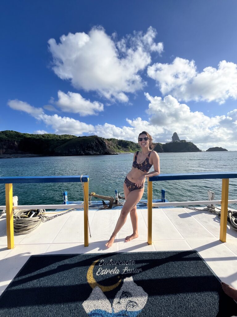 Mulher de biquini sorrindo no convés de um barco com o oceano e ilhas rochosas ao fundo, sob um céu azul.
