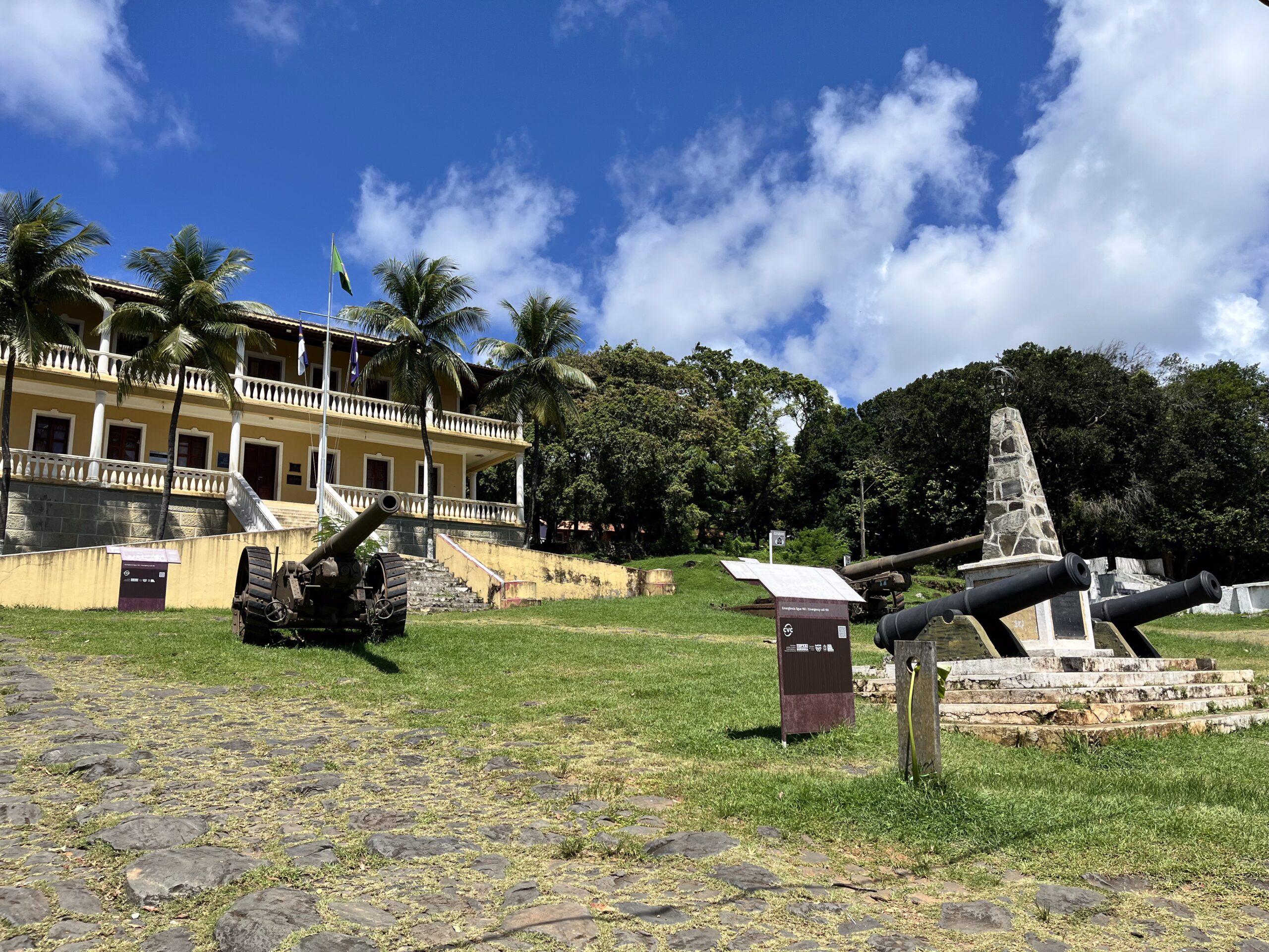Edifício histórico com uma bandeira brasileira, canhões e um monumento de pedra em uma área gramada sob um céu azul com nuvens.