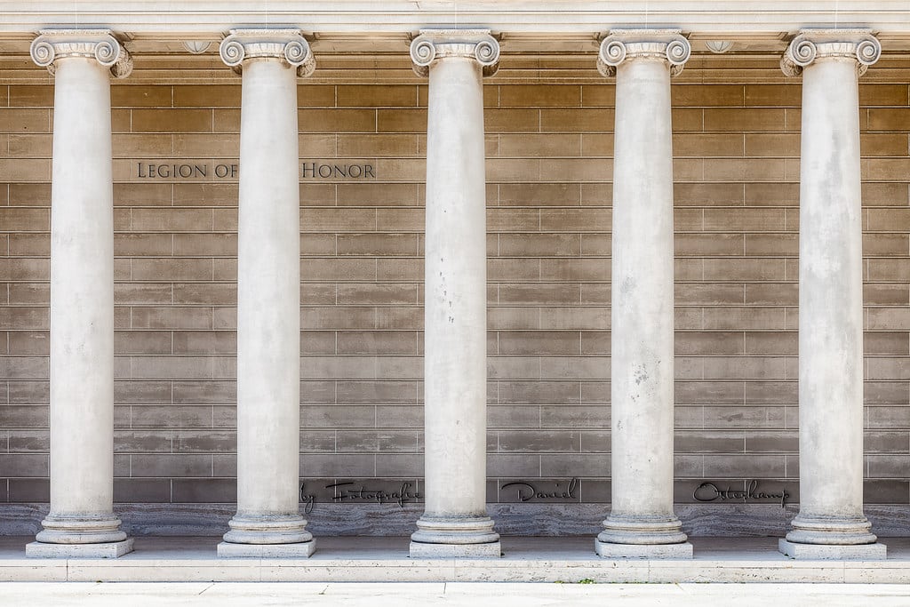Cinco colunas clássicas em frente a uma parede bege. A parede tem o texto "Legion of Honor" e algumas assinaturas perto da base.