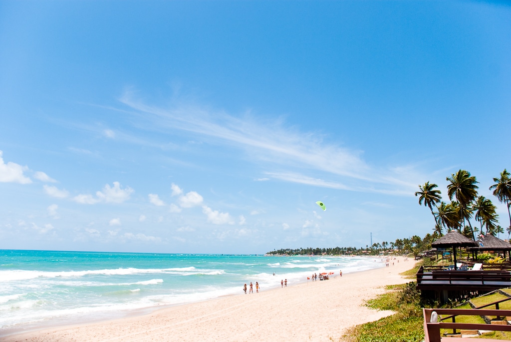 Uma praia de areia com pessoas caminhando ao longo da costa, palmeiras e um parapente no céu sob um céu azul brilhante.
