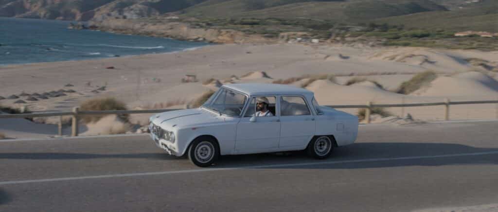 Um carro branco antigo passa por uma estrada costeira com uma praia de areia e penhascos rochosos ao fundo na Praia do Guincho, um dos pontos turísticos de Portugal.