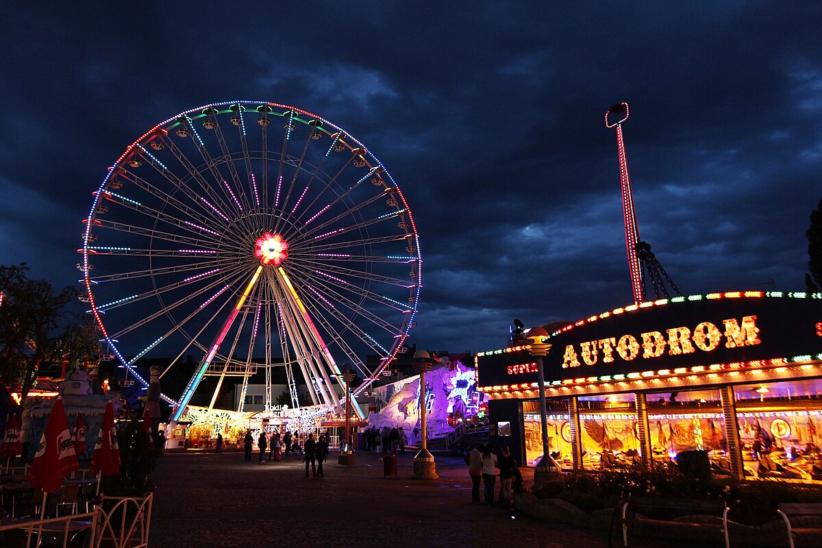 Prater durante à noite noturno com uma roda gigante iluminada, placa "Autodrom" iluminada e pessoas caminhando sob um céu nublado. Essa é uma ótima opção de o que fazer em Viena.