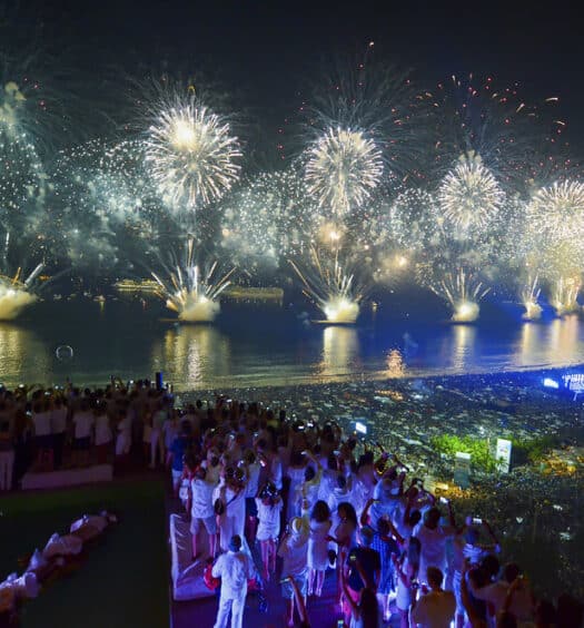 Pessoas vestidas de branco assistem a uma grande exibição de fogos de artifício sobre uma cidade e orla à noite. Essa é a queima de fogos em Copacabana, um dos melhores lugares para curtir o Réveillon no Brasil.