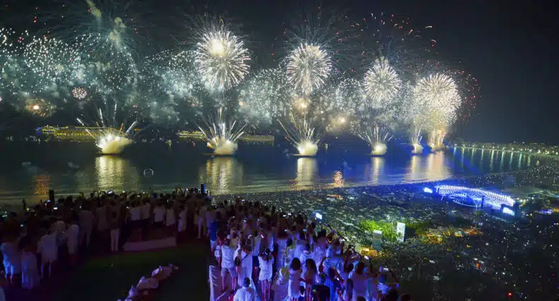 Pessoas vestidas de branco assistem a uma grande exibição de fogos de artifício sobre uma cidade e orla à noite. Essa é a queima de fogos em Copacabana, um dos melhores lugares para curtir o Réveillon no Brasil.